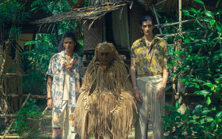 Two male models wearing Kapten Batik printed shirts from the PERSONA 2026 collection, standing alongside a Mah Meri Moyang Bajos mask figure at Pulau Carey