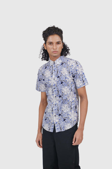 A model wearing a short-sleeve button-up shirt with a blue and white floral batik pattern, paired with black trousers, against a light gray background.